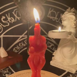 Peut inclure: Une bougie rouge en forme de torse humain est allumée avec une flamme vive. À droite, une figurine de chérubin blanc tient une petite bougie chauffe-plat allumée. L'ensemble est placé sur un fond sombre avec un motif circulaire occulte.