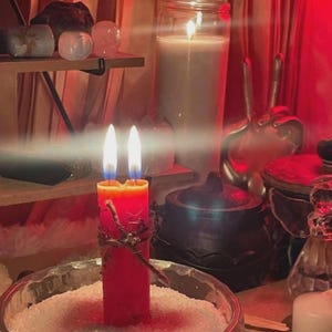May include: A double-wick red pillar candle wrapped in twine, standing upright in a clear glass bowl filled with white granular salt. The candle has two bright flames and is positioned in front of a large white candle and a black cauldron.