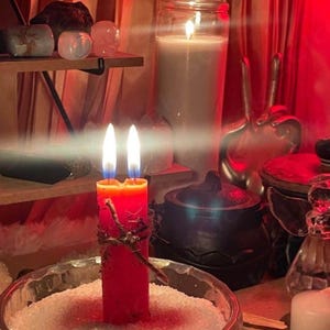 May include: A red candle with two flames, decorated with a twig, sits in a glass bowl filled with white crystals. Other candles, crystals, and ritual items are visible in the background, creating a mystical atmosphere. The scene is illuminated with a warm, reddish glow.