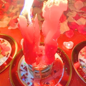 Peut inclure: Deux grandes bougies rose vif brûlant dans un bocal en verre, avec de la cire fondue coulant sur les côtés. Les bougies sont placées sur une assiette entourée de pétales de rose rouges et blancs, avec une petite bougie chauffe-plat en forme de cœur.