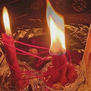 May include: Two red wax candles standing in silver foil bases, each topped with a bright flame. A thin red string is wrapped around both candles, connecting them in a web-like pattern. The wax appears textured and uneven, with visible melting.