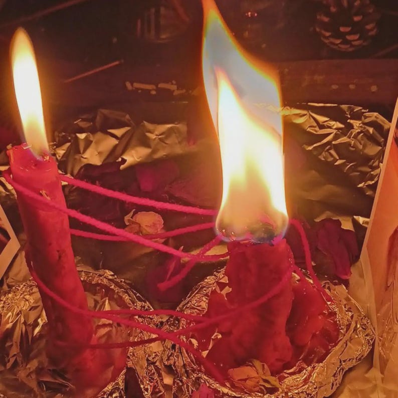 May include: Two red wax candles standing in silver foil bases, each topped with a bright flame. A thin red string is wrapped around both candles, connecting them in a web-like pattern. The wax appears textured and uneven, with visible melting.
