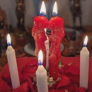 Könnte beinhalten: Eine Ansammlung brennender Kerzen, darunter zwei große rote Kerzen, die mit roter Schnur zusammengebunden sind. Drei weiße Kerzen sind ebenfalls angezündet, mit sichtbaren Flammen. Die Kerzen sind auf einer roten Oberfläche mit roten Rosenblättern angeordnet.