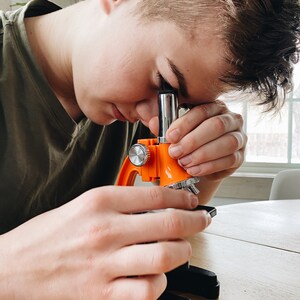 May include: A person looking through the eyepiece of an orange and silver microscope. The microscope is on a wooden table.
