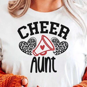May include: White t-shirt with black text that reads "Cheer Aunt" with a red heart and a black and white leopard print heart on either side of a white megaphone.