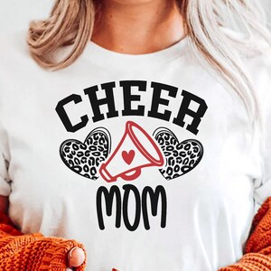 Könnte beinhalten: Weißes T-Shirt mit dem Text "CHEER MOM" in schwarzen Buchstaben. Der Text ist von zwei roten Herzen mit einem schwarzen Leopardenmuster umgeben. Ein rotes Herz befindet sich in einem weißen Megaphon.