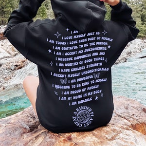 May include: Black hooded sweatshirt with a purple print of affirmations and a graphic of a globe with the text "A Kinder Planet" on the back. The text includes phrases like "I love myself just as I am today", "I am capable", and "I accept myself unconditionally".