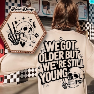 Può includere: Una felpa color crema con la scritta "WE GOT OLDER BUT WE'RE STILL YOUNG" e una grafica a teschio. Un design a tasca con un teschio e un segno di pace è a sinistra. Lo sfondo presenta un ristorante.