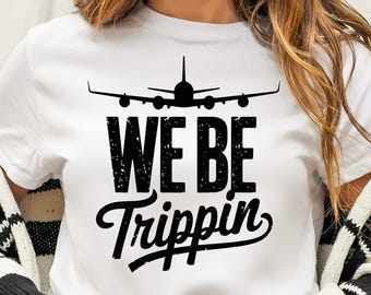 Op de afbeelding: Wit T-shirt met een zwart silhouet van een vliegtuig boven de woorden "WE BE Trippin" in een versleten lettertype. Het shirt heeft een ronde hals en lijkt van een zacht materiaal te zijn.