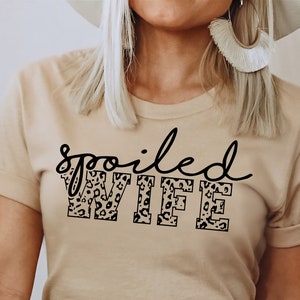 May include: A beige t-shirt with the text "spoiled WIFE" in black lettering. The letters are filled with a leopard print pattern.