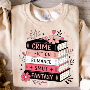 May include: A beige sweatshirt with a stack of books printed on the front. The books are labeled "CRIME", "FICTION", "ROMANCE", "SMUT", and "FANTASY". The sweatshirt is decorated with pink flowers.