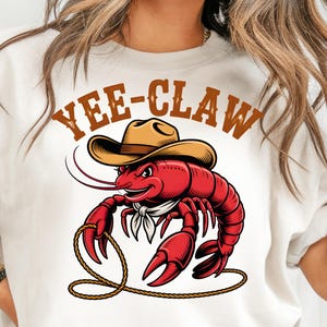 Może przedstawiać: Biała koszulka z kreskówkową ilustracją czerwonego raka w kapeluszu kowbojskim i chuście. Tekst "YEE-CLAW" jest wydrukowany nad rakiem brązowymi literami. Rak trzyma linę.