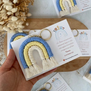 May include: A blue, yellow and white macrame rainbow keychain with a gold key ring. The keychain is attached to a white card with text in Italian that reads: "L'Amore è una percezione intimamente legata ai colori, come migliaia di arcobaleni sovrapposti uno sull'altro. Thomas 22 OTTOBRE 2029 Macramegiftaccessory"