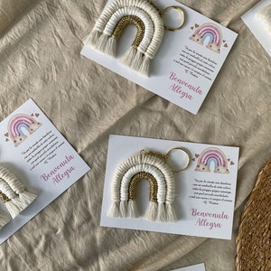 May include: A set of three white and gold rainbow keychains with a small card attached to each. The card has a pink and blue rainbow design and the text "Va per la strada una bambina con un ombrella di sette colori sotto la pioggia grigia cammina con quel piccolo arcobaleno e nel suo cuore è sempre il serena Benvenuta Allegra".