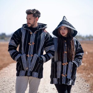 Op de afbeelding: Twee jacks met capuchon voorzien van een zwart-wit geweven patroon met verticale strepen en geometrische details. Elk jack heeft een puntige capuchon, lange mouwen, twee voorzakken en drie houten knevelsluitingen aan de voorzijde.