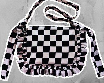 May include: A rectangular shoulder bag featuring a black and white checkered pattern. The bag includes a matching checkered shoulder strap and decorative ruffled fabric trim along the sides and bottom edges of the main compartment.