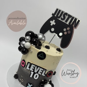 May include: A gaming-themed cake with a cylindrical shape, featuring a gradient from grey to cream. It has a black controller topper with the name "Justin" and "Level 10" text. Decorated with black, silver, and white spheres, and gaming symbols.