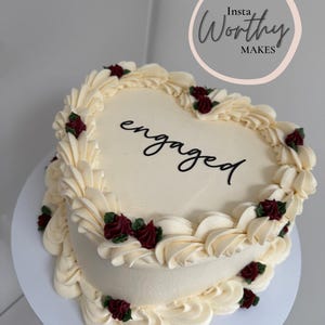 May include: A heart-shaped cake decorated with white icing and red roses. The cake is inscribed with the word "engaged" in black lettering. The cake is on a white cake stand.