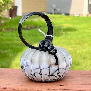 May include: A hand-blown glass pumpkin sculpture featuring a white and translucent body with dark blue webbed detailing. The piece is topped with a tall, black, spiraled glass stem that curves into a circular loop at the very top.