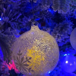 May include: A close-up of a Christmas tree ornament. The clear glass ball is covered in glitter and features a snowflake design. The ornament hangs from a silver string and is surrounded by blue and green Christmas lights.