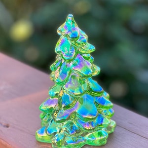 May include: A decorative glass Christmas tree with a green iridescent finish. The tree has a layered design with a pointed top and a rounded base. The glass reflects shades of green, blue, and purple, creating a shimmering effect. The tree is approximately 5 inches tall.