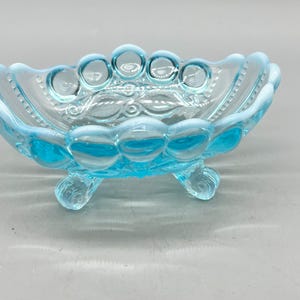 May include: A light blue glass bowl with a scalloped rim and a repeating circular pattern along the sides. The bowl features three small, curved feet at the base and an embossed swirl design on the interior floor, crafted from translucent glass.