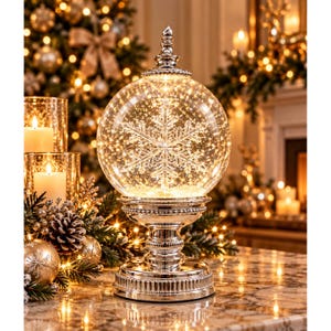 May include: A clear glass globe ornament featuring an internal illuminated snowflake design, mounted on a polished silver-toned pedestal base with a decorative finial top, emitting a warm glow from integrated internal lights.