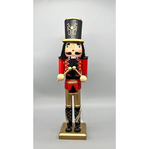 May include: A wooden nutcracker figurine dressed as a drummer, featuring a black top hat with gold scrollwork, a red jacket with gold accents, gold trousers, and black boots. The figure holds two gold drumsticks over a red and black drum.