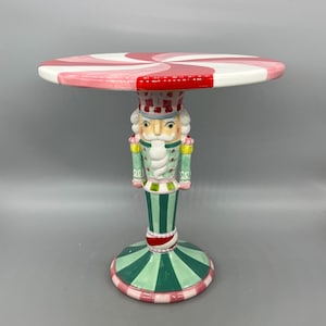 May include: A whimsical, decorative side table featuring a candy cane-inspired top and a nutcracker figure base. The round tabletop has a red and white spiral design. The base is green, red, and pink, with a nutcracker figure in the center.