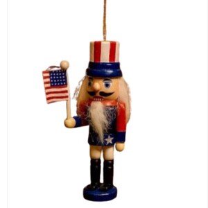 May include: A wooden Uncle Sam nutcracker ornament, painted in red, white, and blue. The figure holds a small American flag and has a long white beard. The ornament is suspended by a gold string.