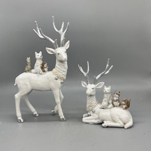 May include: Two white deer figurines with silver antlers and hooves. One deer stands while the other reclines. Both feature small woodland animals, including a fox, owl, and squirrel, perched on their backs. Each deer wears a decorative collar.