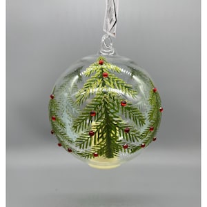 May include: Clear glass Christmas ornament with a green glitter Christmas tree design and small red bead accents. The ornament is round and has a loop for hanging. A festive holiday decoration.