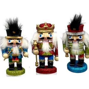 May include: Three decorative nutcrackers with glittery bases. One nutcracker is in a blue uniform with a black hat and plays the drums. Another wears a red and gold crown and holds a staff. The third nutcracker is in a green uniform and holds a sword.