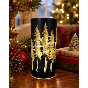 May include: A cylindrical black lantern featuring a cut-out design of evergreen trees. The interior is illuminated by warm white fairy lights, which shine through the tree silhouettes, casting a golden glow against the dark exterior surface.