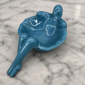 May include: A glossy, teal-colored sculpture of a seated figure. The figure is in a relaxed pose, with legs extended and arms wrapped around the body. The sculpture has a smooth, reflective surface and is likely made of ceramic or glass.