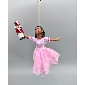 May include: A hanging ornament featuring a ballerina with dark hair wearing a pink dress with glittery accents. The ballerina holds a small nutcracker figure in one hand. The ornament is suspended by a thin gold cord for display purposes.