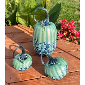 May include: Three glass pumpkins in varying sizes, featuring a pale mint green iridescent finish with vertical ribbing and dark blue speckled patterns at the base. Each pumpkin is topped with a curved, dark blue glass stem with a spiral detail.