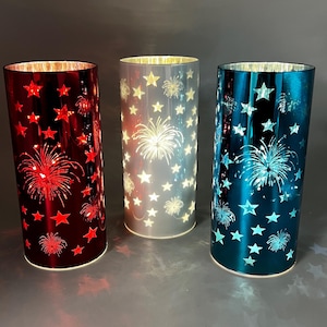 May include: Three cylindrical candle holders in red, silver, and teal. Each has a pattern of stars and fireworks cut into the sides. The interior glows with light, creating a festive ambiance.