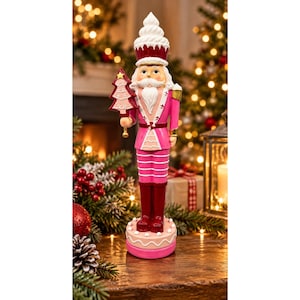 May include: A nutcracker figurine wearing a pink and dark red uniform with a white cupcake-shaped hat. The figure holds a pink Christmas tree ornament and stands on a round base decorated with white icing patterns and small dots.