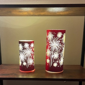 May include: Two cylindrical red decorative lamps with a fireworks design. The lamps are illuminated from within, creating a warm glow. The lamps are different sizes and are placed on a wooden shelf.