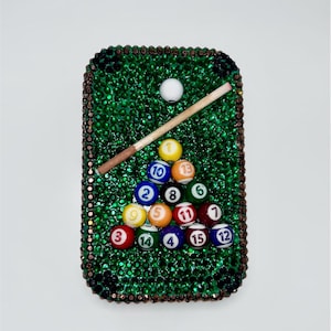 May include: A rectangular phone case covered in green rhinestones resembling a pool table. It features a miniature white cue ball, a wooden pool cue, and a triangular rack of fifteen numbered billiard balls in various colors including yellow, blue, red, and orange.
