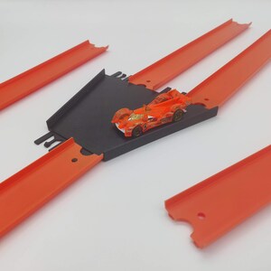 May include: A toy car race track set with orange plastic track pieces and a black plastic ramp. The track is assembled in a cross shape with a toy car on the ramp.