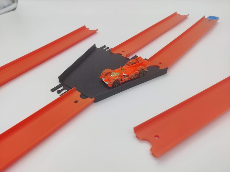 May include: A toy car race track set with orange plastic track pieces and a black plastic ramp. The track is assembled in a cross shape with a toy car on the ramp.