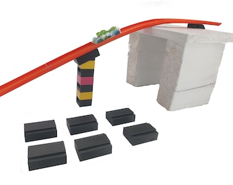 May include: A toy car racing down an orange track, elevated by a multi-colored support structure. The track leads to a white wooden platform. Several black blocks are arranged at the base, suggesting a construction or play set.