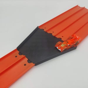 May include: A red plastic toy race track with a black plastic ramp and a small orange toy car on the ramp.