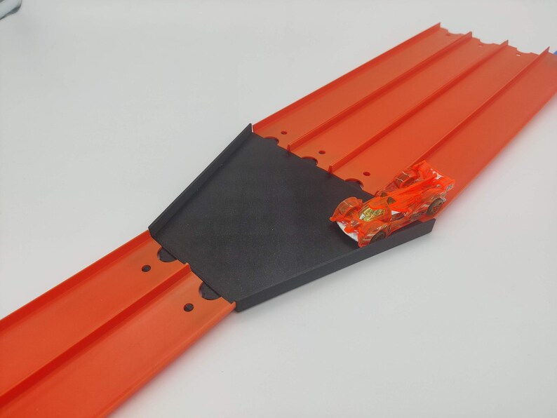 May include: A red plastic toy race track with a black plastic ramp and a small orange toy car on the ramp.