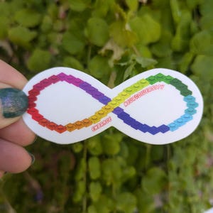 Puede incluir: Una pegatina blanca con un símbolo de infinito hecho de ladrillos de Lego de colores. El texto "EMBRACE NEURODIVERSITY" está escrito en la pegatina.