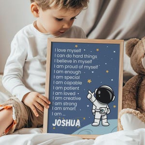 Może przedstawiać: Oprawiony wydruk z niebieskim tłem, ilustracją astronauty i imieniem "JOSHUA". Wydruk zawiera pozytywne afirmacje, takie jak "Kocham siebie" i "Jestem silny". Rama jest jasnobrązowa.