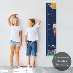 May include: A navy blue height chart with a space theme. The chart features a rocket, astronaut, and a sun with the name "Noah". The chart measures up to 6 feet. Two children stand next to the chart.
