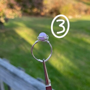 K&ouml;nnte beinhalten: Ein Ring aus silbernem Draht mit einem hellvioletten Edelstein. Der Ring wird von einer Zange gehalten.